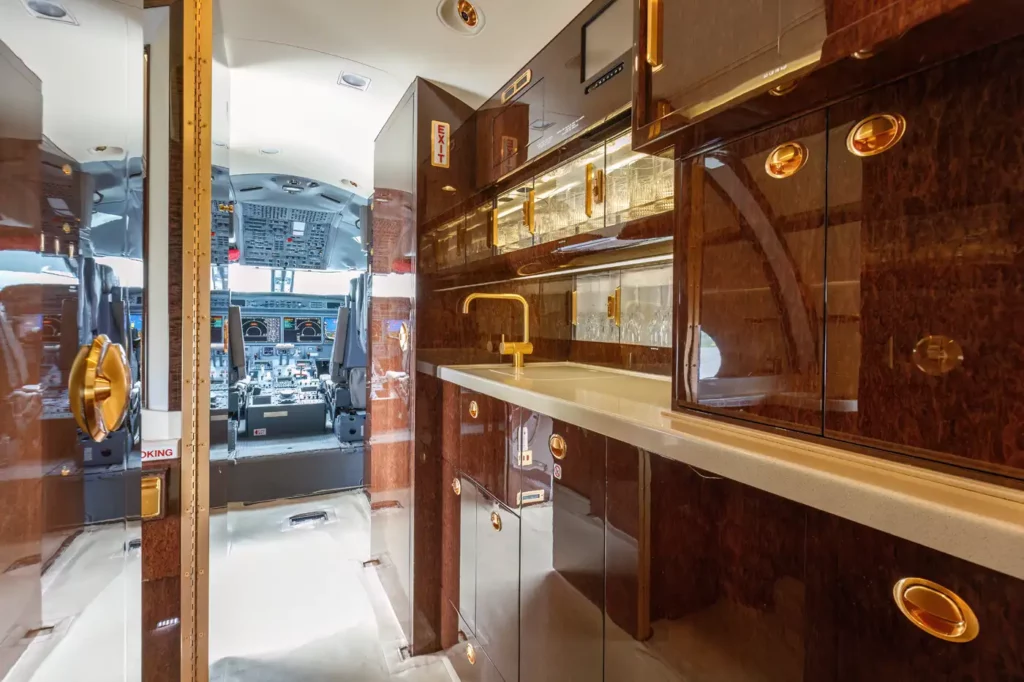 Galley area of the Gulfstream G550, equipped with elegant wood finishes, gold fixtures, and a display of crystal glassware, with the cockpit visible in the background.