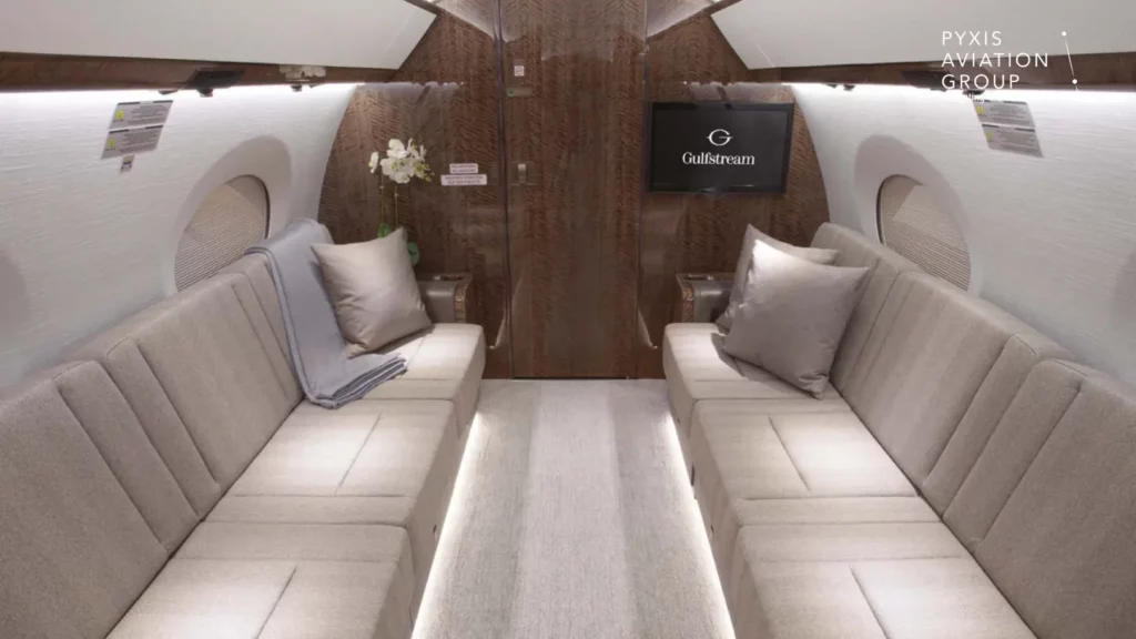 Interior of a Gulfstream G650 jet, showing the aft cabin with two long, beige sofas facing each other. The cabin is furnished with soft cushions and blankets, and features polished wood paneling, ambient lighting, and a small side table with a plant and decorative accents.