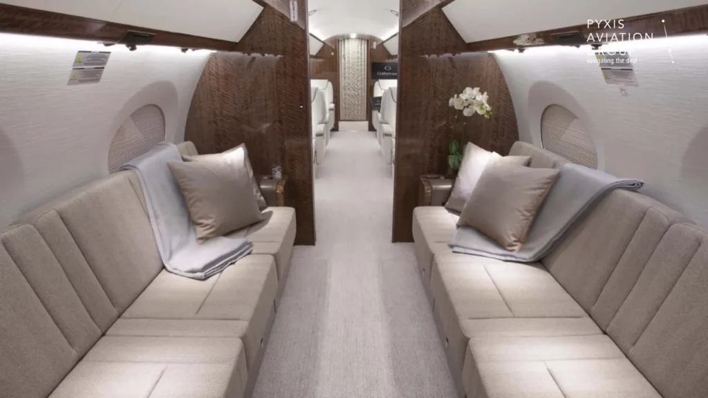 Interior of a Gulfstream G650 jet, featuring a modern, luxurious cabin with light beige sofas arranged along both sides. The seating area includes plush cushions and blankets, with the cabin decorated in elegant wood paneling and soft lighting.