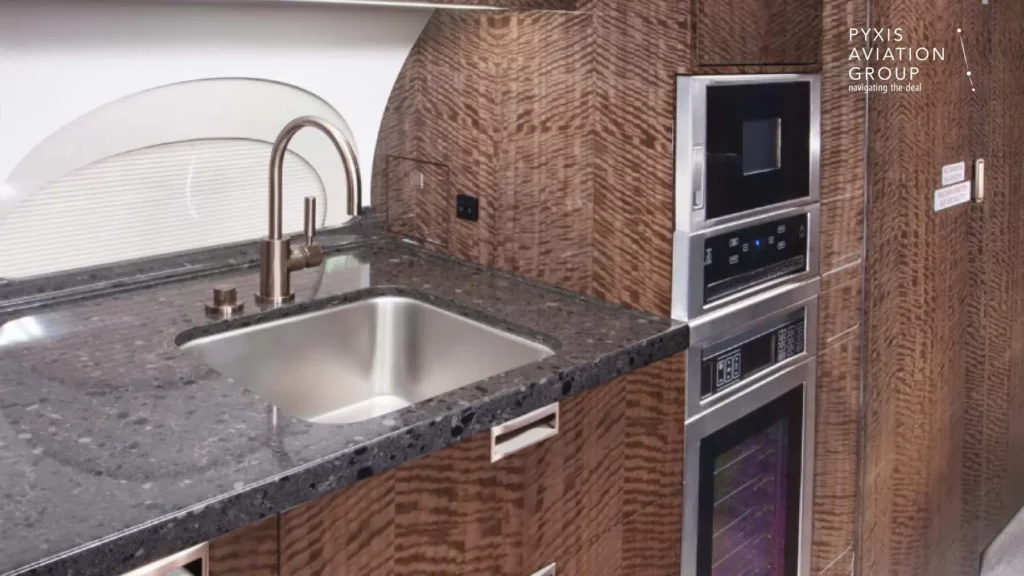 Galley area of a Gulfstream G650 jet, featuring a modern kitchenette with a stainless steel sink, polished countertops, and built-in appliances. The galley is adorned with rich wood cabinetry, providing a high-end, functional space for in-flight meal preparation.