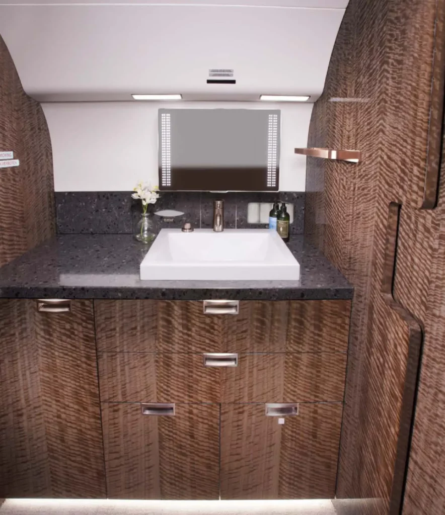 Lavatory sink area in a Gulfstream G650 jet, showcasing a sleek, modern design with rich wood cabinetry, a dark stone countertop, and a rectangular white sink. Above the sink is a large mirror with built-in lighting, and the space is accented with a small vase of flowers and luxury toiletries.