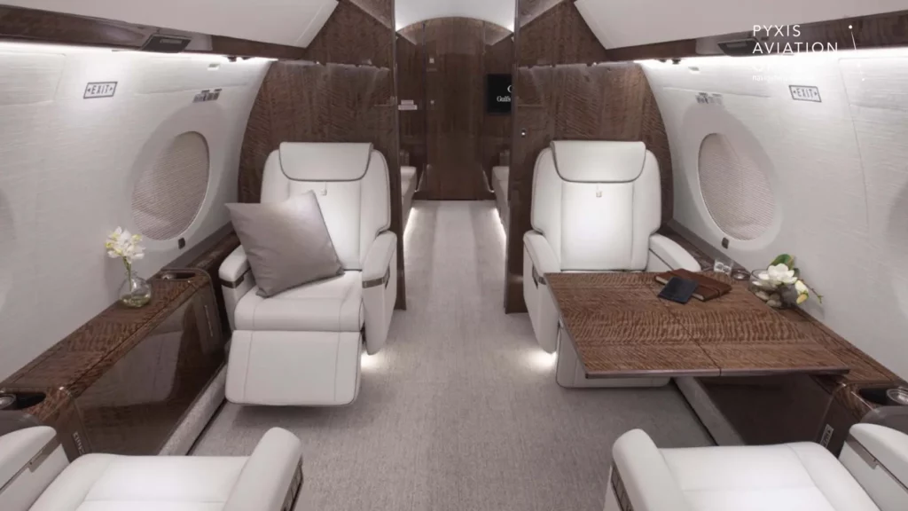 Mid-cabin of a Gulfstream G650 jet, featuring two plush white leather seats with a wooden table between them. The table is set with a few personal items, including a smartphone, notebook, and a small vase with flowers, adding a touch of elegance and practicality to the luxurious seating area.