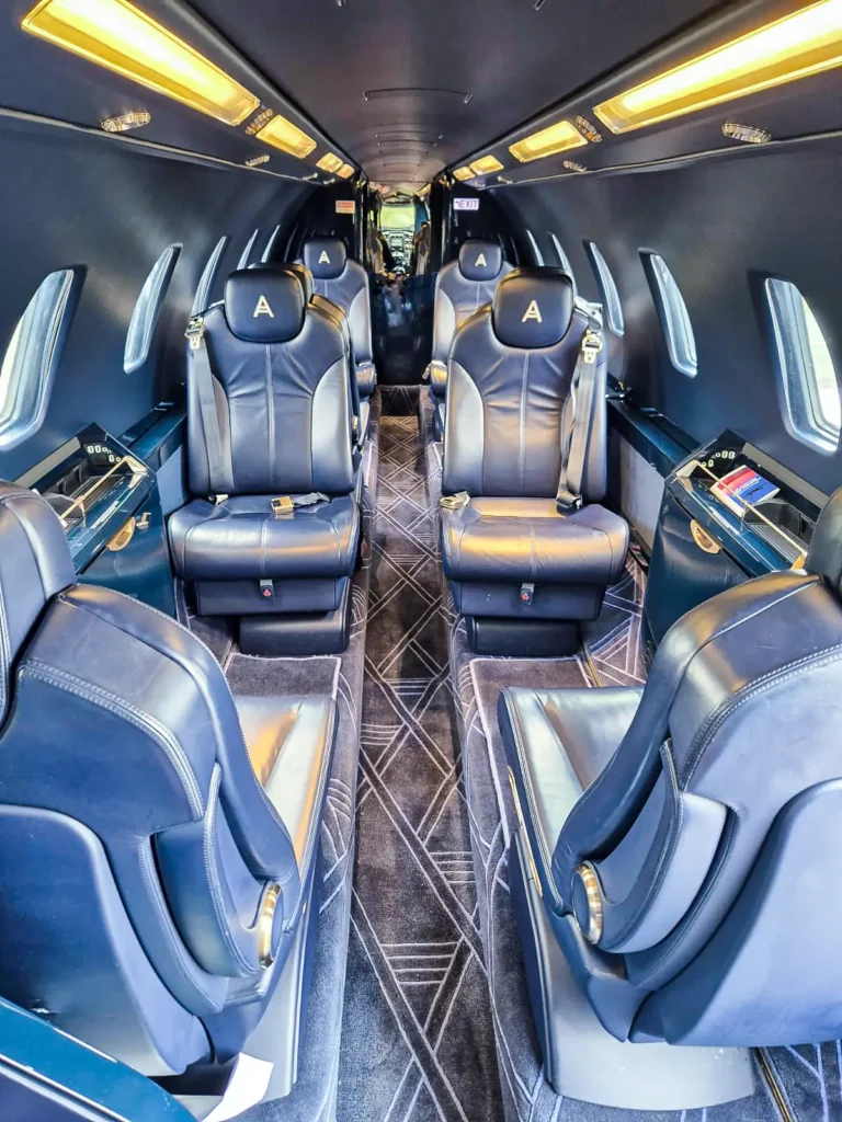 The luxurious interior of a Cessna Citation Sovereign+ private jet featuring plush leather seats with embroidered "A" logos. The seating is arranged in pairs along the sides of the cabin, with ample legroom and wood-panel accents. Overhead lighting illuminates the space, and windows line the walls, providing natural light.