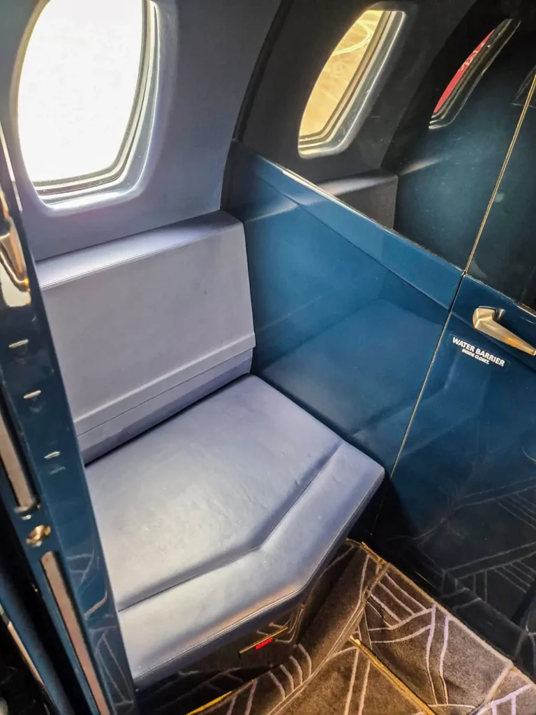 The sleek lavatory in a Cessna Citation Sovereign+, featuring a cushioned seat and two small windows allowing natural light. The design is minimalist with soft blue upholstery and a small storage compartment next to the seat.