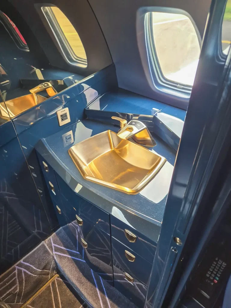 The well-designed lavatory sink area of a Cessna Citation Sovereign+ with a geometric-shaped metal sink and a modern faucet. It sits on a glossy blue counter with multiple storage drawers beneath, surrounded by small windows for natural light.